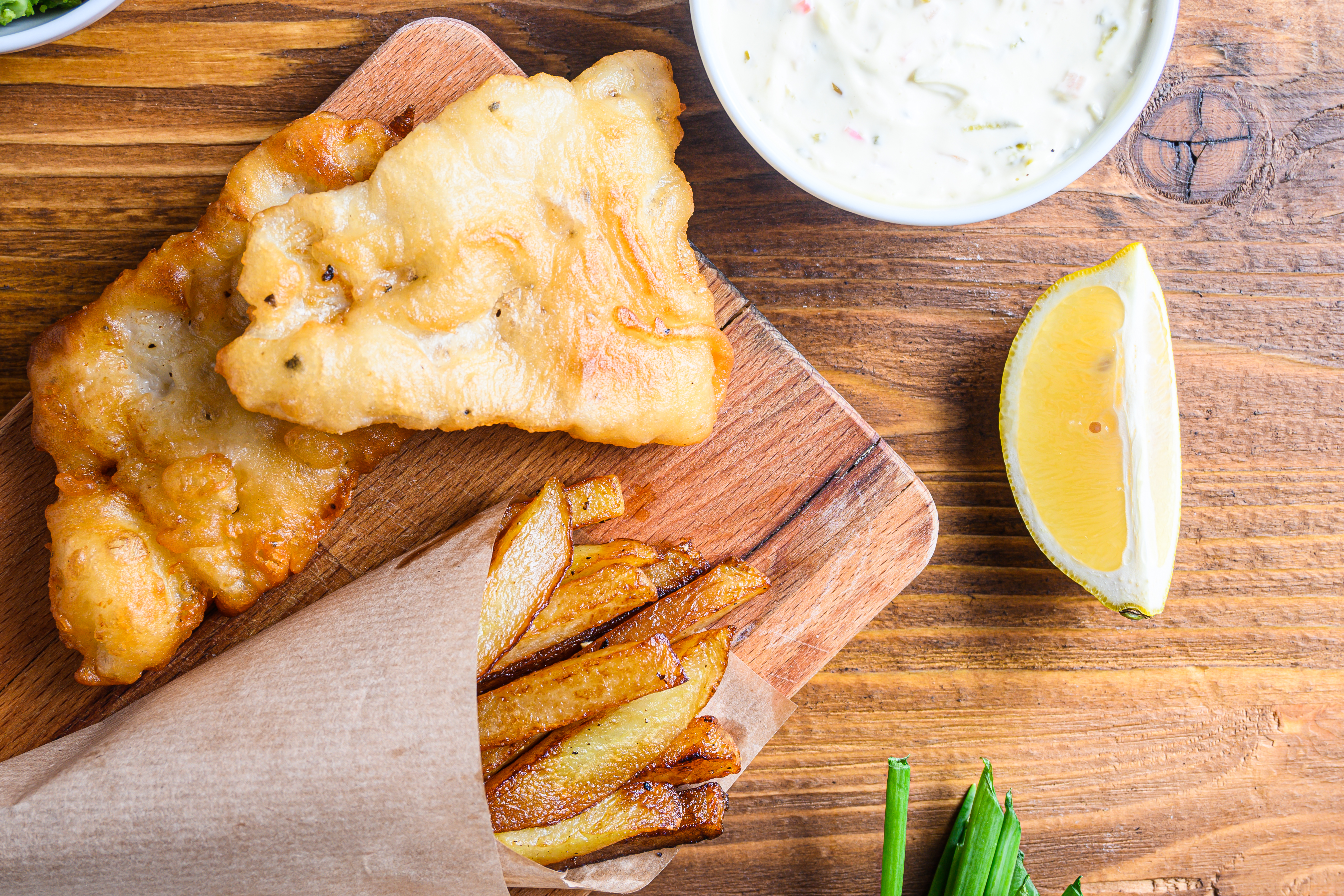 Detail of fish chips with dip and lemon, mashed minty peas, tartar sauce in paper cone  on wood chopping board dip and lemon - fried cod, french fries, lemon slices, tartar sauce, ketchup tomatoe served in the Pub or  over light old wooden planks table top view close up.