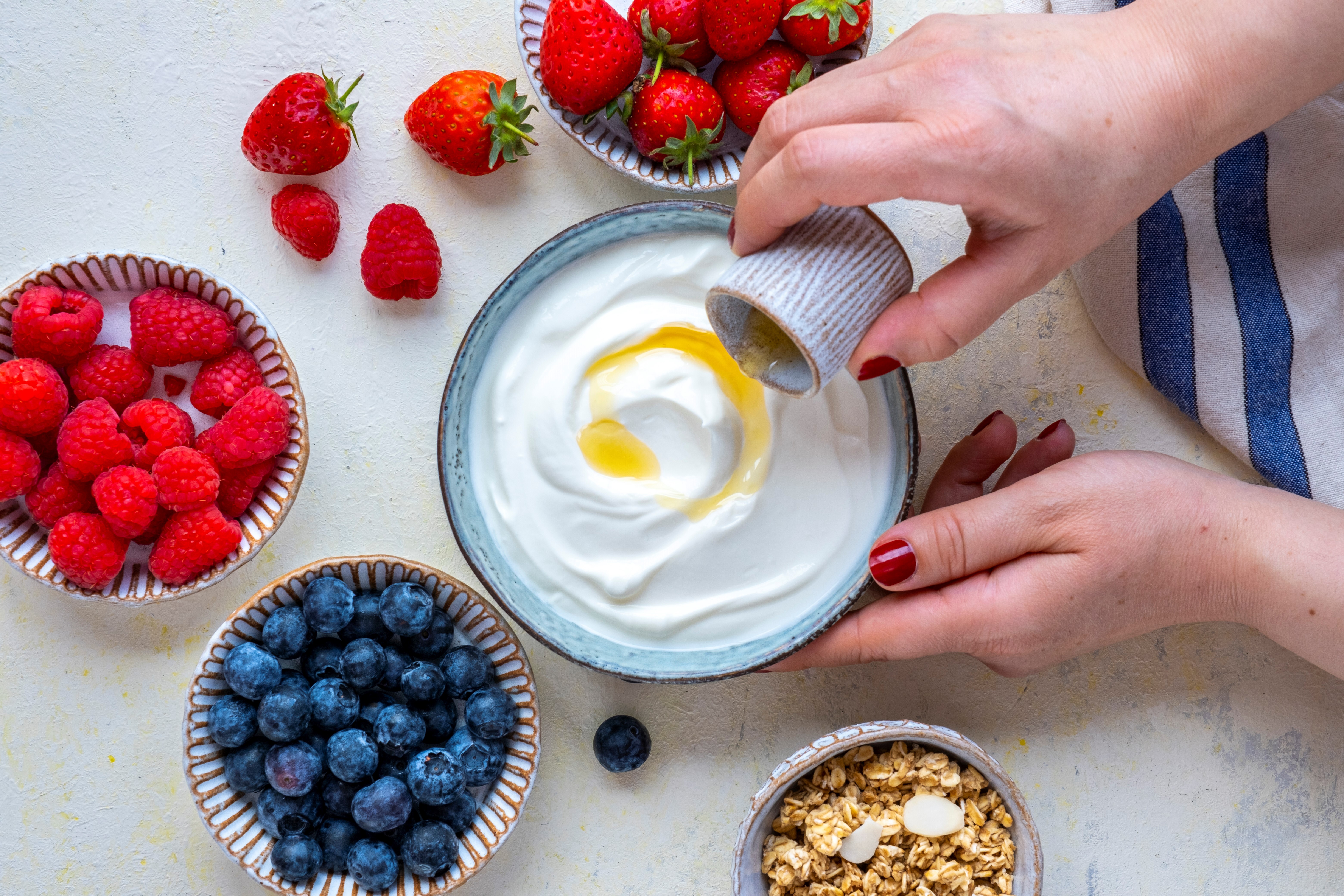 Woman hands pouring maple syrup over Greek yogurt, raspberries, strawberries, blueberries and granola on the side.