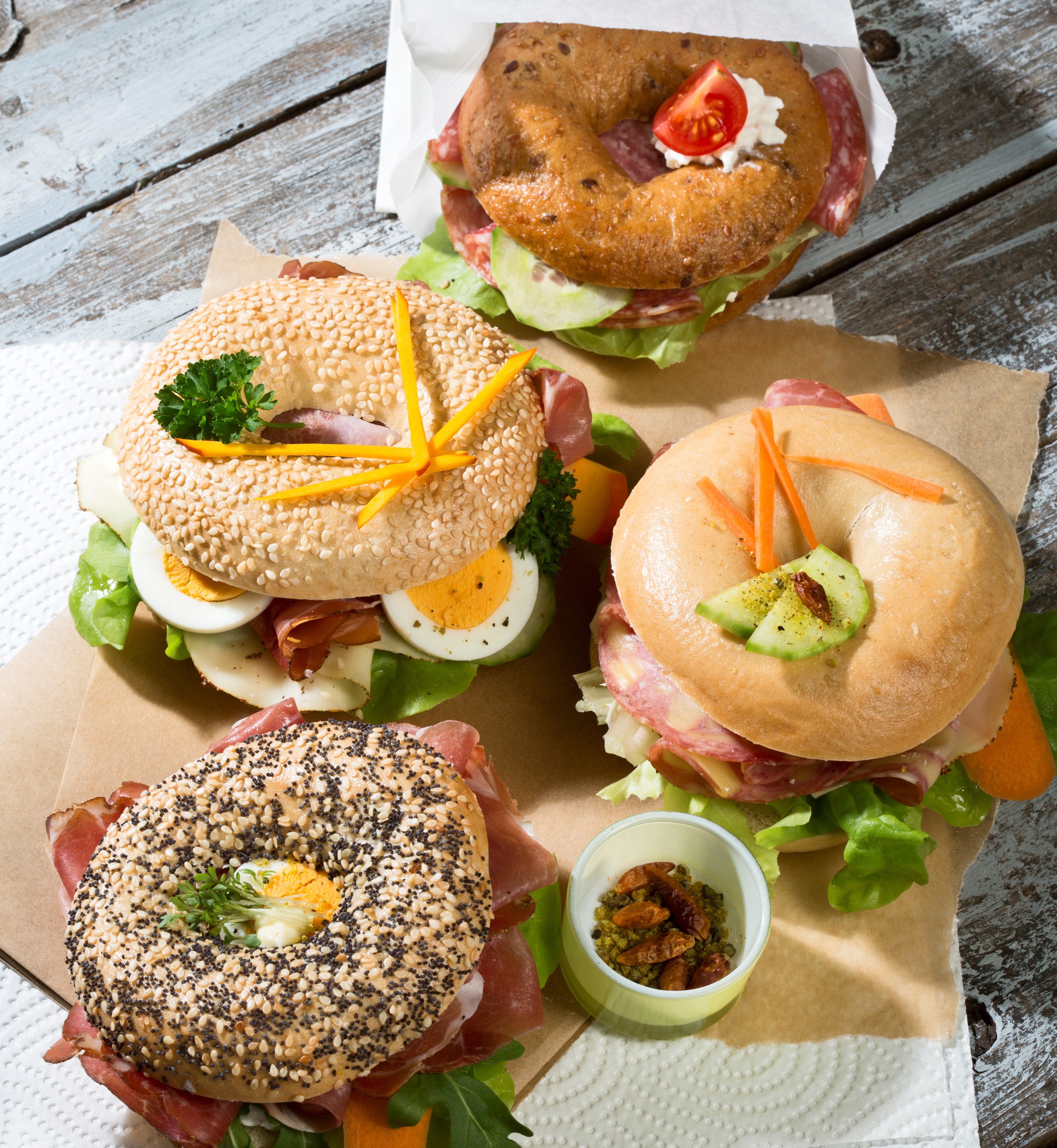 Four different bagels garnished with salami, sausage, slices of bacon, rocket salad, tomato, lettuce, cucumber carrot, egg, cream cheese and cress and parsley