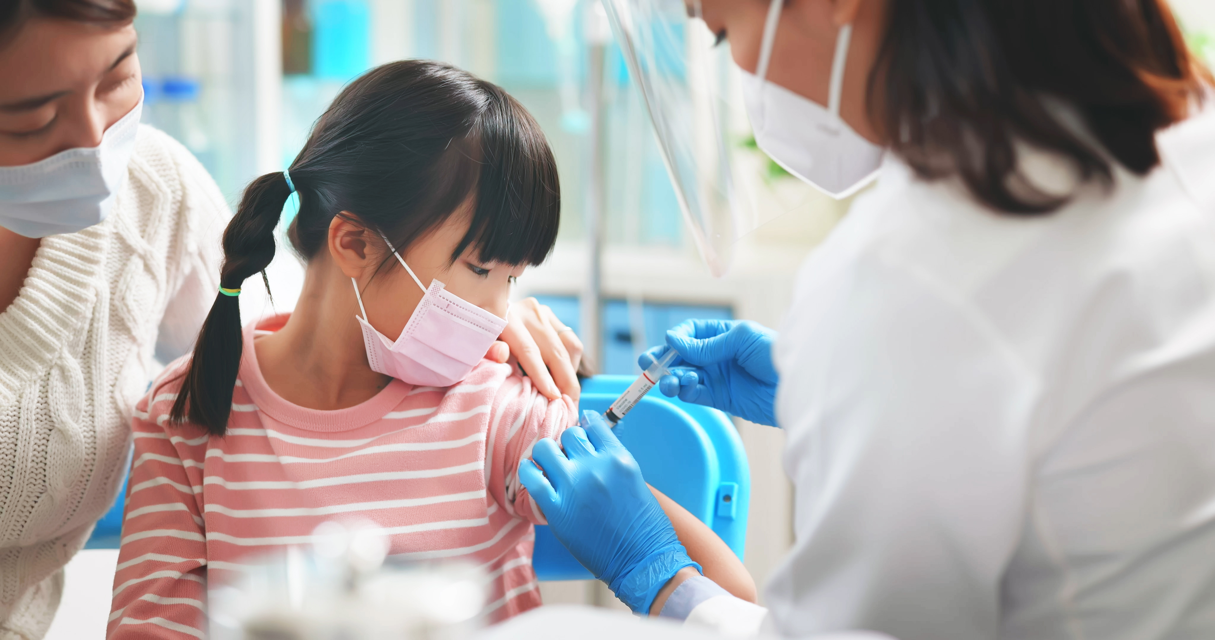 asian female doctor wearing gloves and isolation mask is making a COVID-19 vaccination in the shoulder of child patient with her mother at hospital