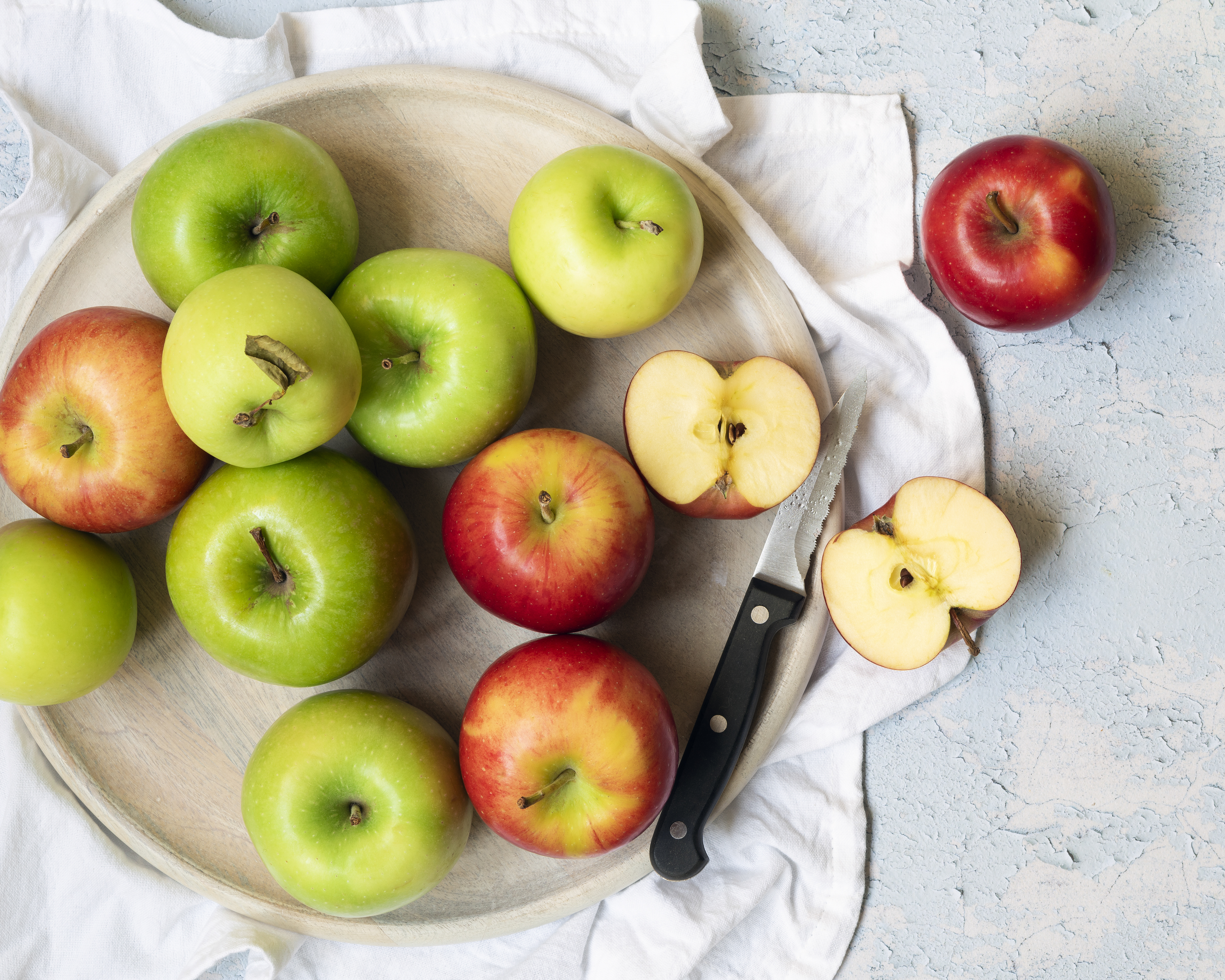 Green and red apples with two apple halves and knife on a round wooden serving board.