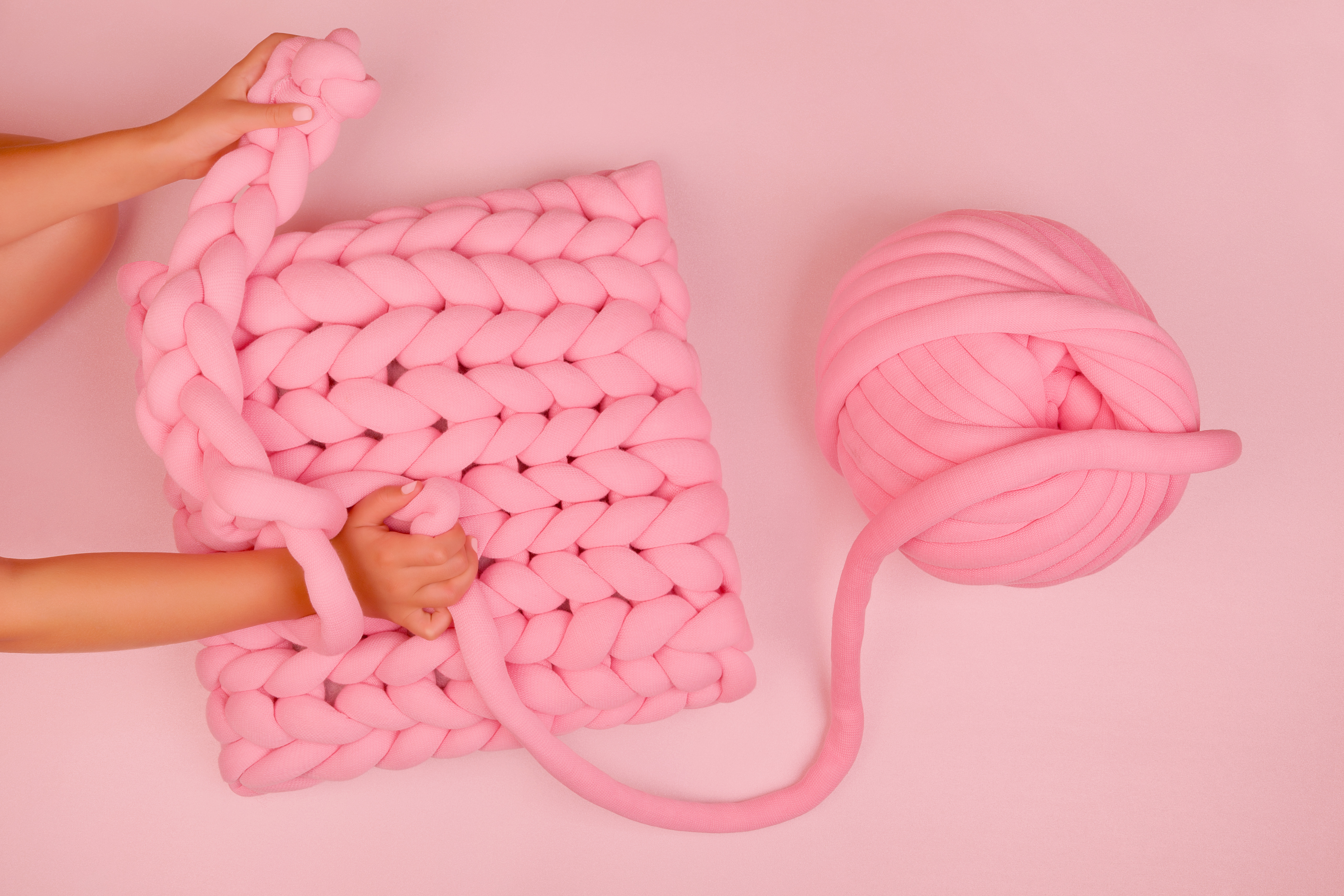 Girl knitting adorable super chunky pillow. Minimalistic pink background. Craft, handmade and skill.
