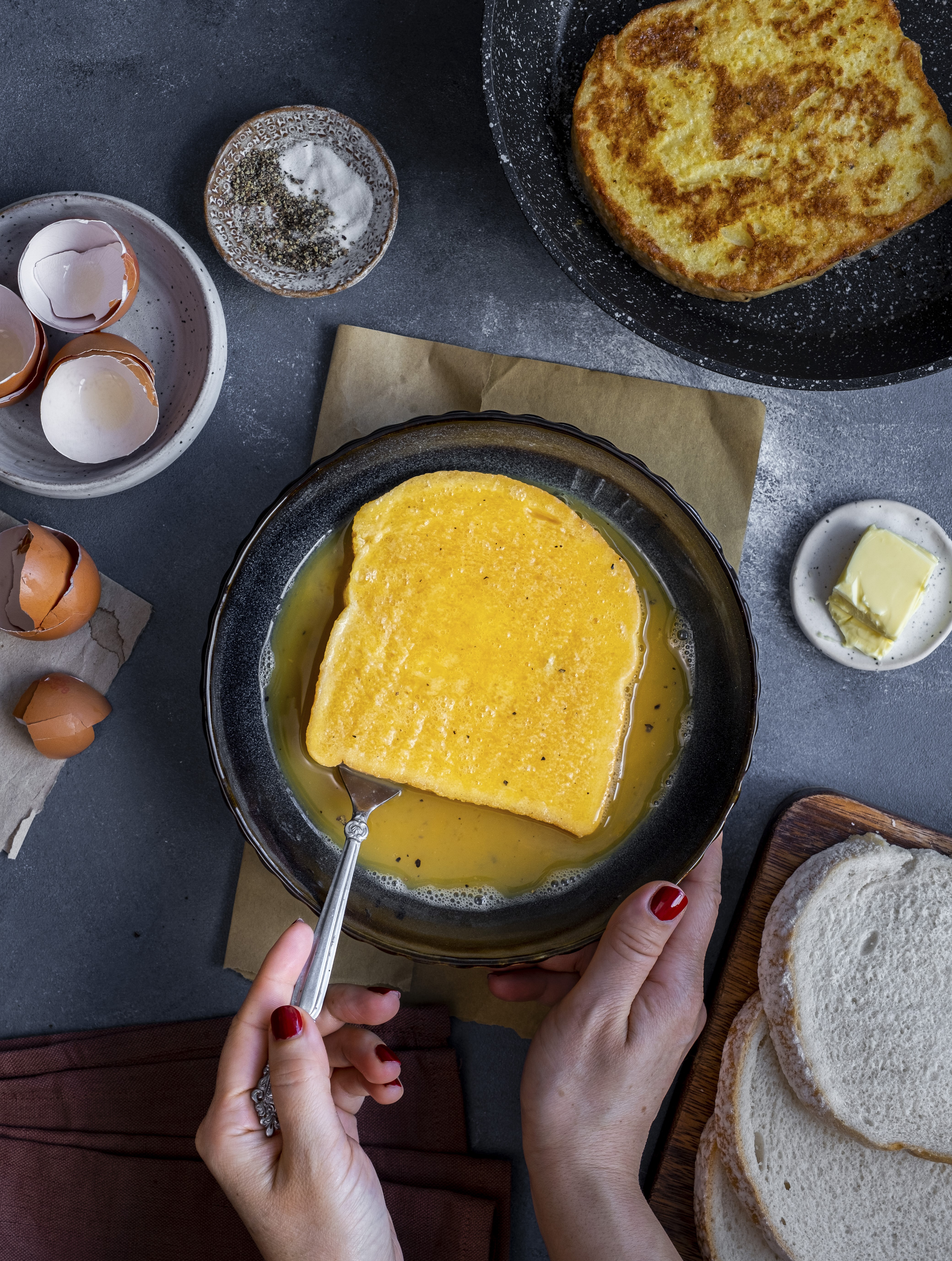 A slice of white bread being soaked in the egg and milk mixture in a bowl and one slice of French toast on a plate on the side.