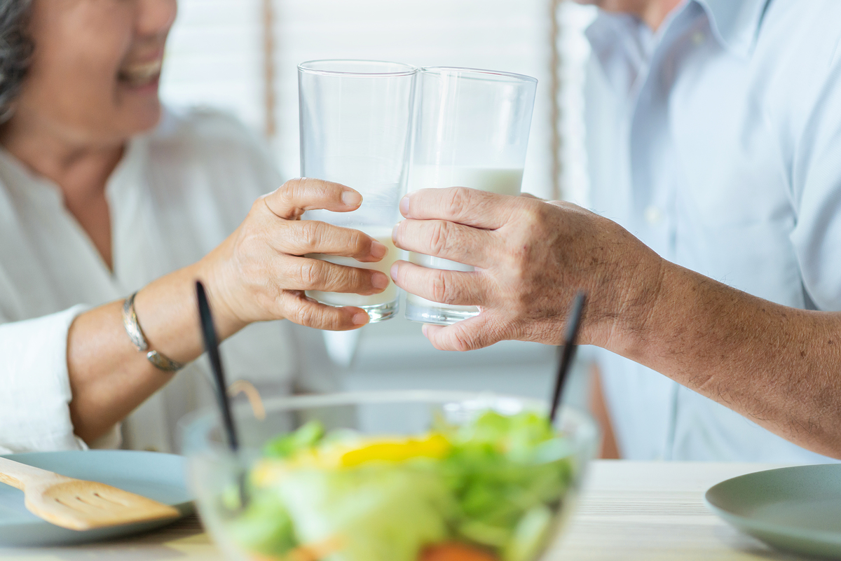 Smiling Asian senior man and woman holding glasses of milk. Cheerful Elderly couple enjoying healthy salad food while having breakfast at home. Happy Lifestyle Grandfather and Grandmother.
